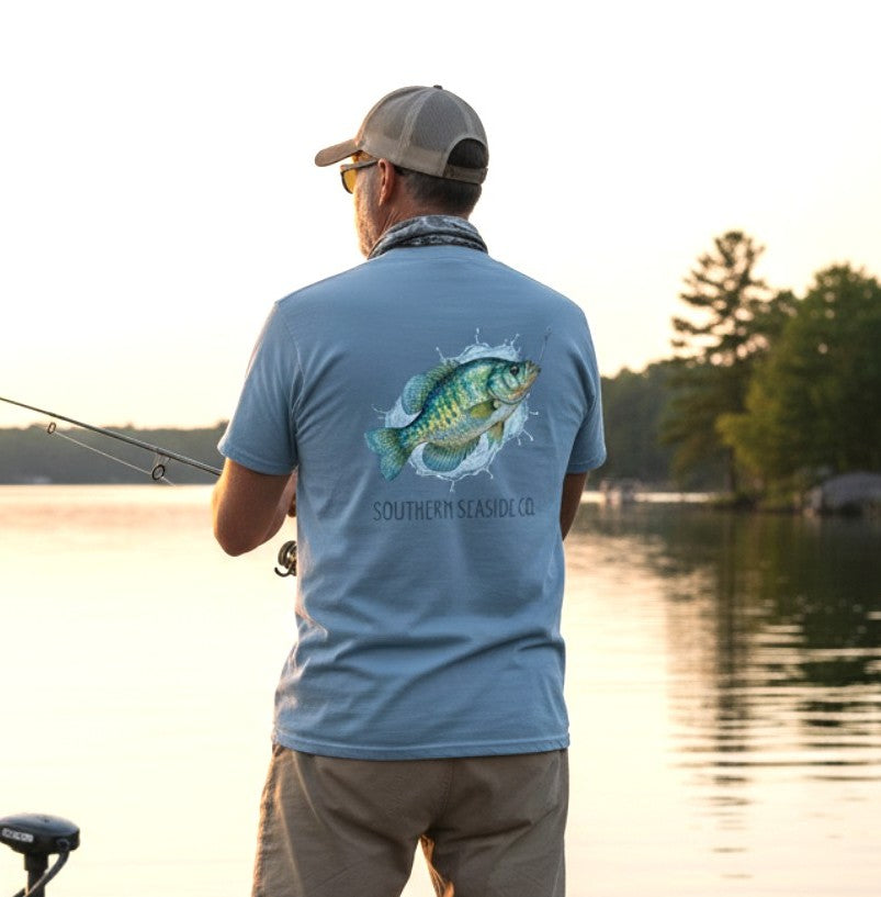 Crappie Fishing T-shirt. A aan fishing by a lake wearing a blue comfort colors t-shirt with a crappie fish design and 'Southern Seaside Co.' text.