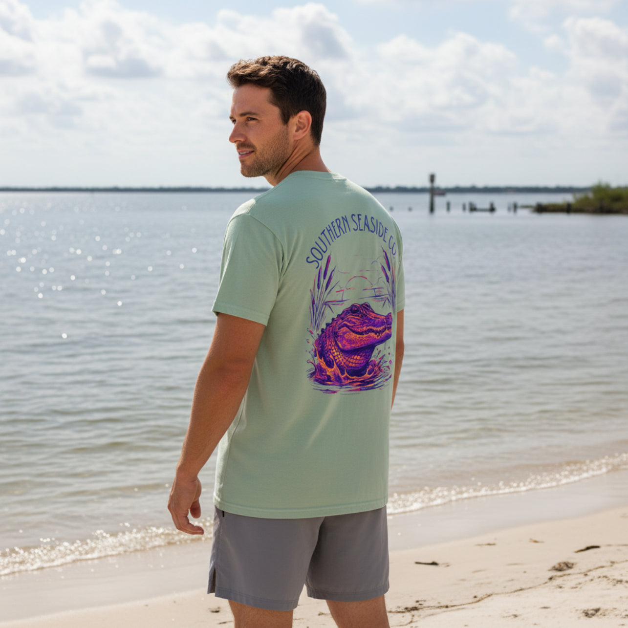 Man wearing a light green t-shirt with an alligator graphic and 'Southern Seaside Co ' text by a body of water.