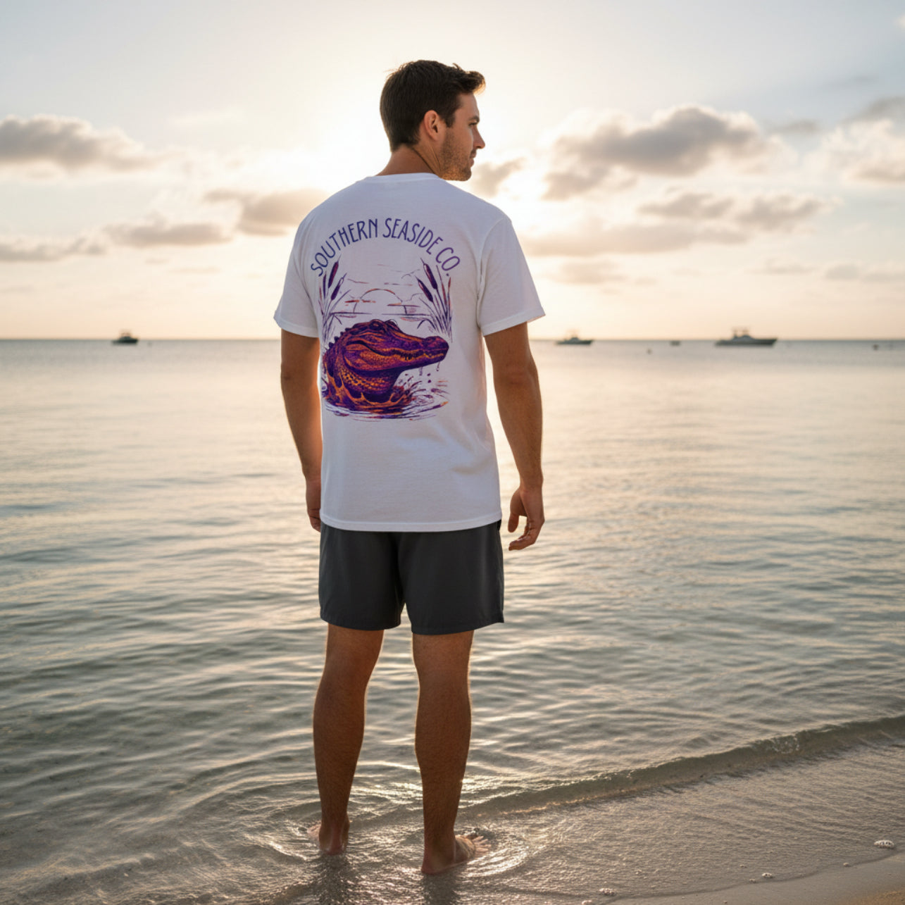 Man standing on a beach wearing a white t-shirt with a colorful alligator design with Southern Seaside logo