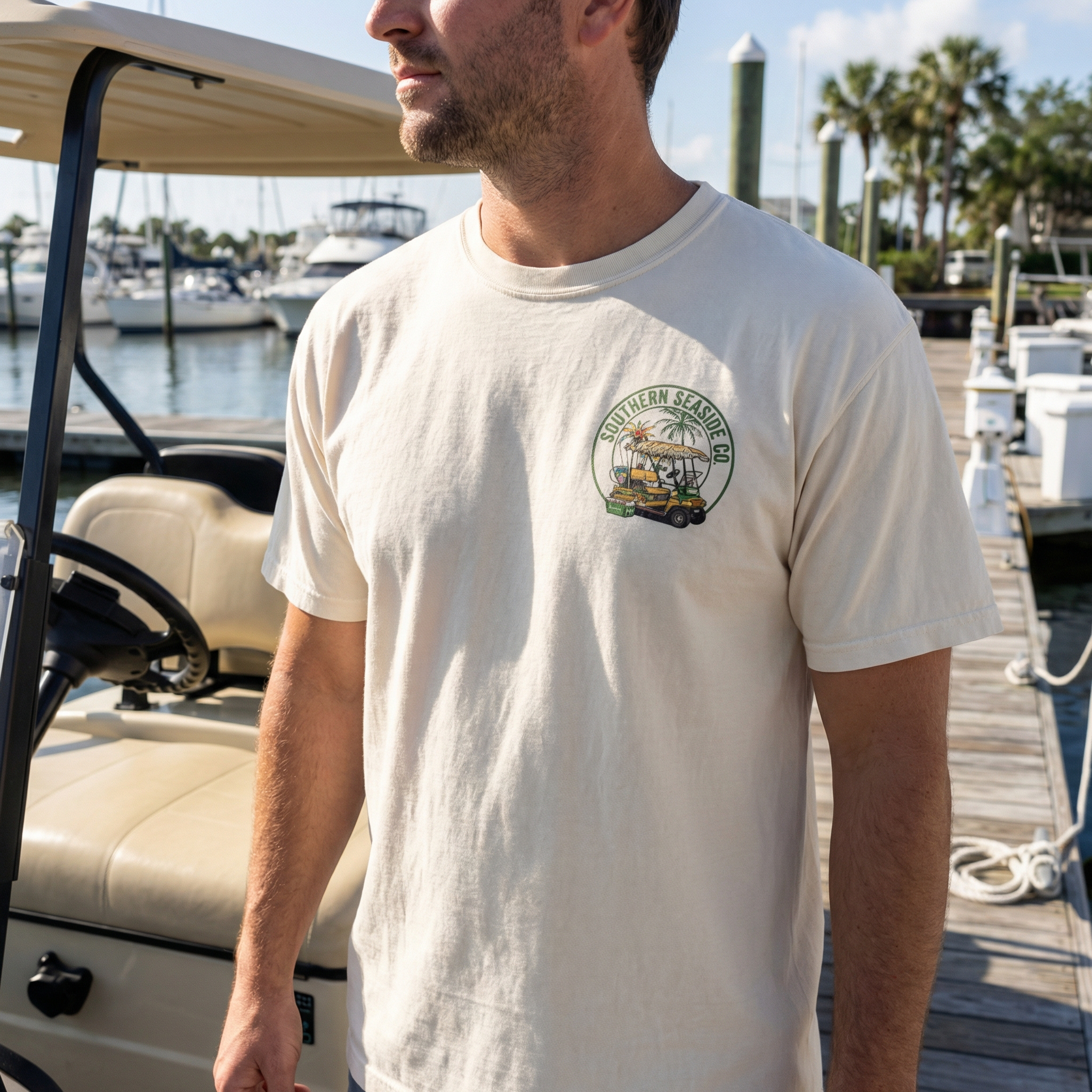 Man wearing a ivory t-shirt with a golf cart and fishing gear logo on a dock with boats in the background