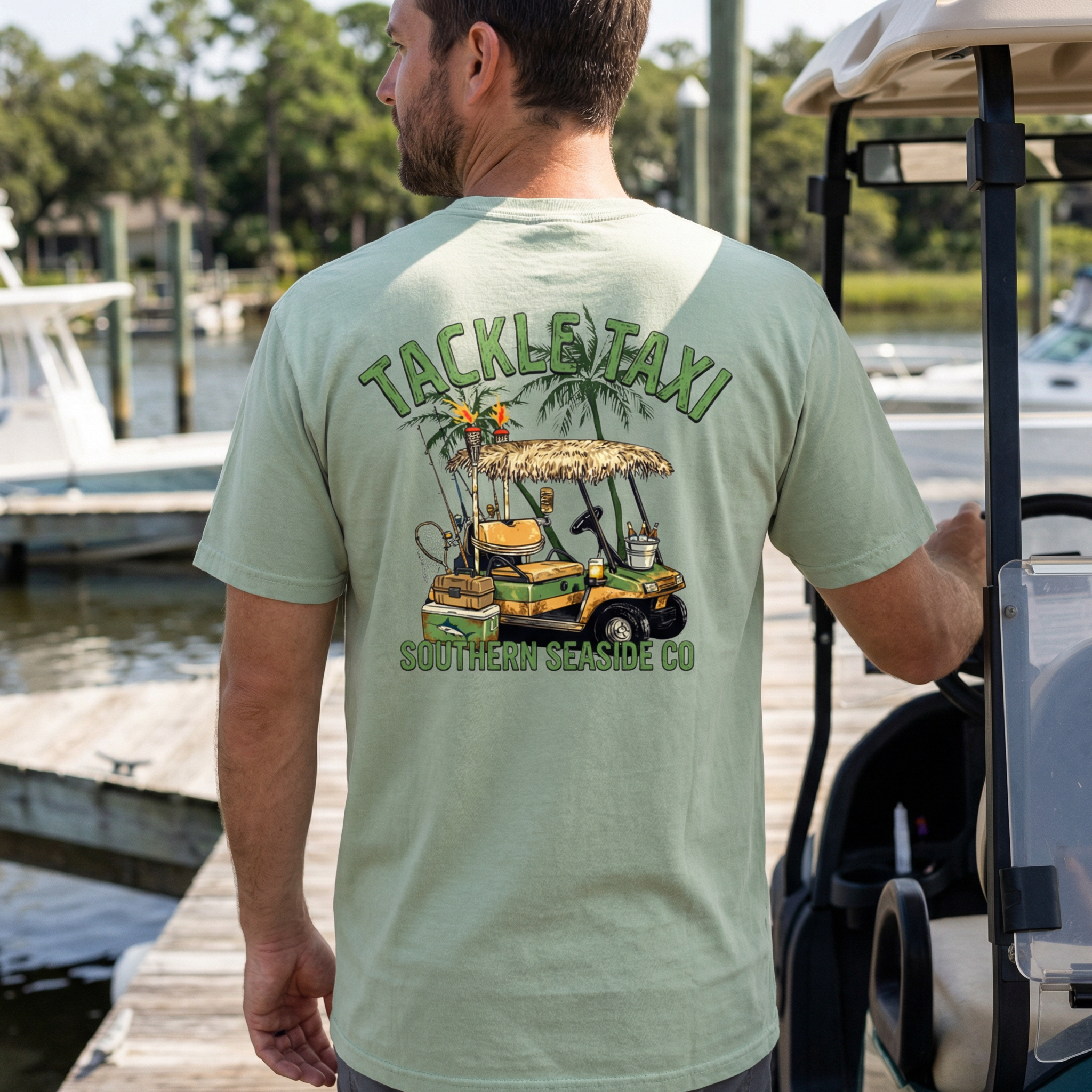 Man wearing a light green t-shirt with a golf cart and fishing gear with 'Tackle Taxi' design by Southern Seaside Co. at a dock.