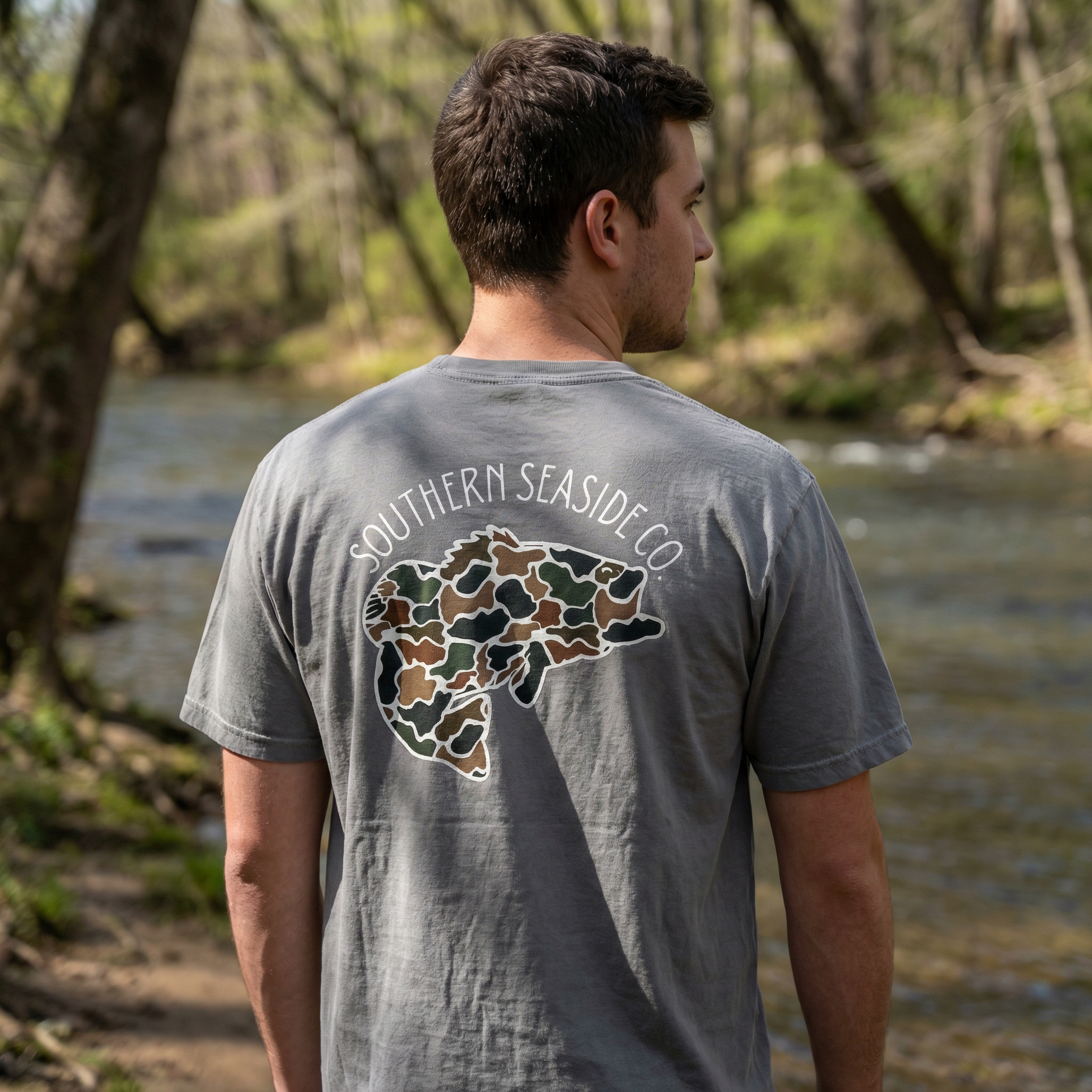 Man wearing a gray t-shirt with bass fish in a camo print pattern and the 'Southern Seaside Co' logo in a forest setting