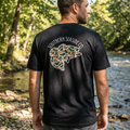 Man wearing a black t-shirt with a camo print bass fish and the 'Southern Seaside Co.' logo outdoors by a river
