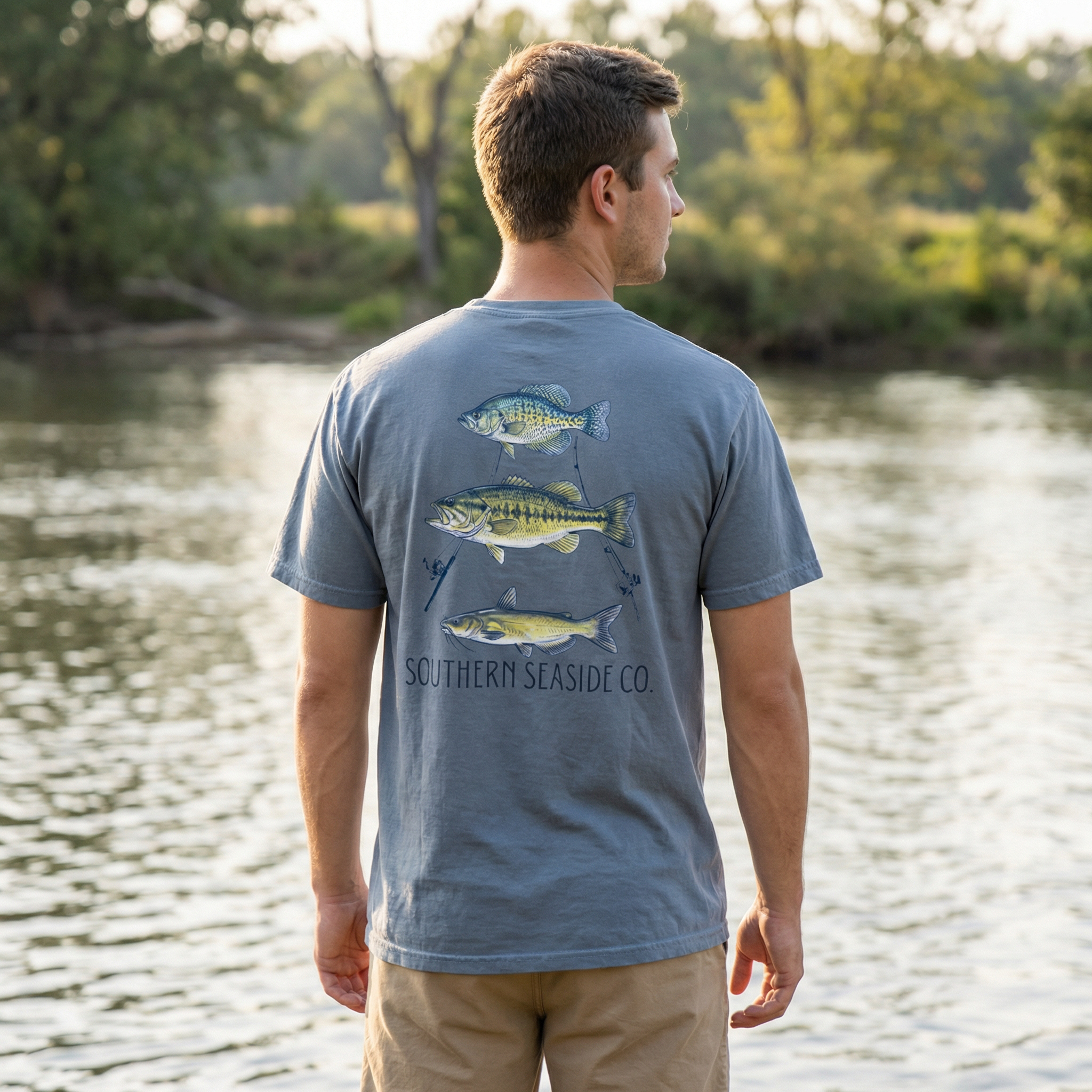 A crappie fish, bass fish and catfish tshirt. Man wearing a blue t-shirt with fish graphics by a body of water