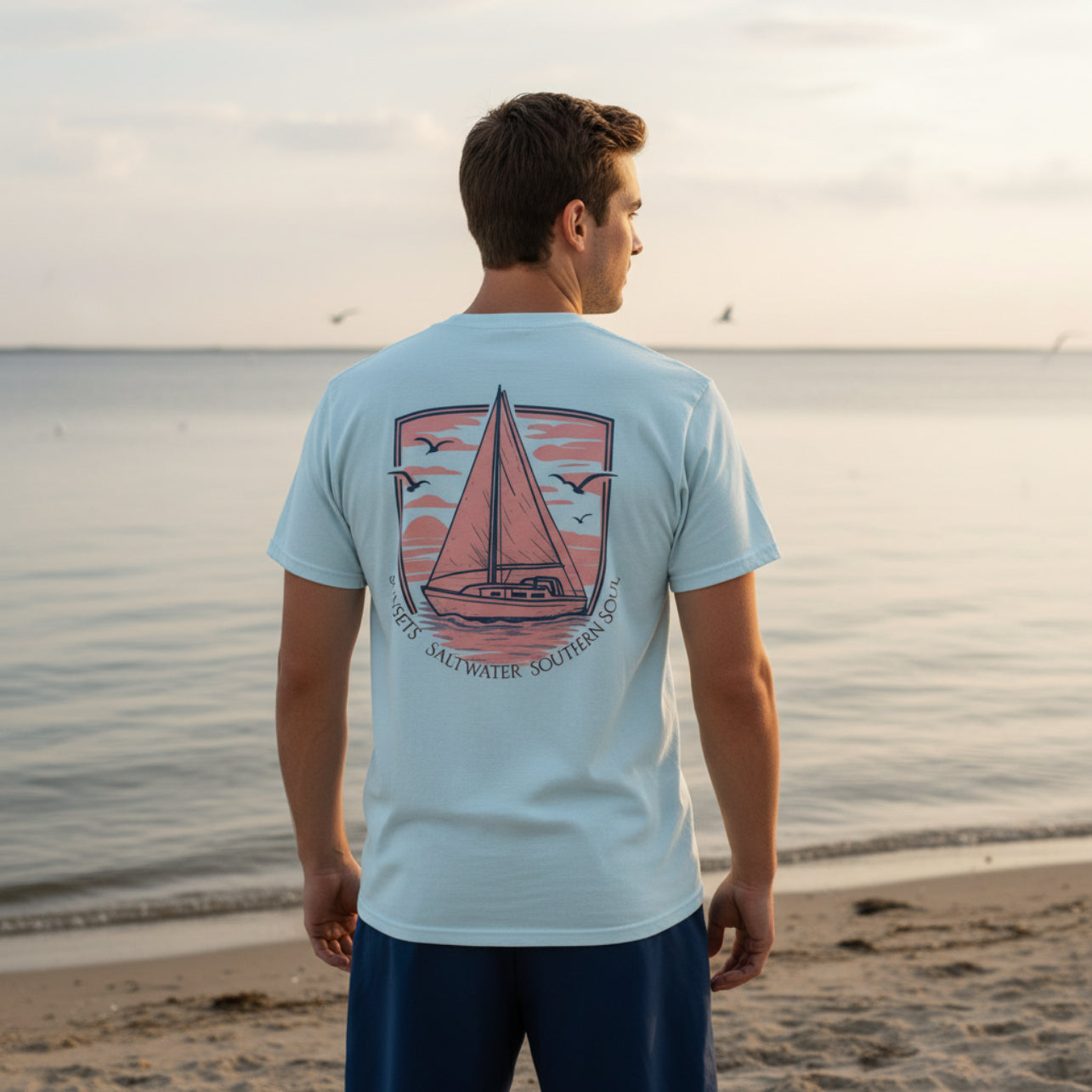 Man wearing a light blue t-shirt with a sailboat design on a beach at sunset.