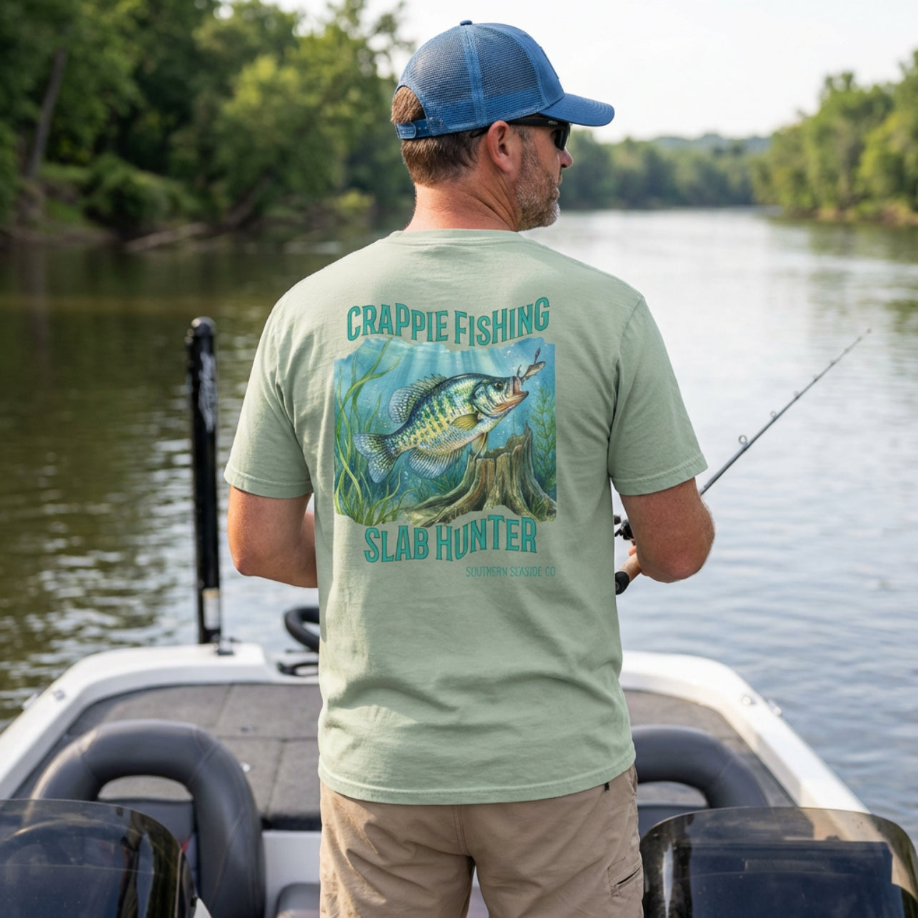 Man on a boat wearing a green t-shirt with crappie fishing design, standing by a body of water.