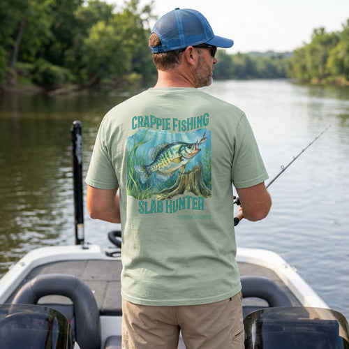 Man on a boat wearing a green t-shirt with crappie fishing design, standing by a body of water.