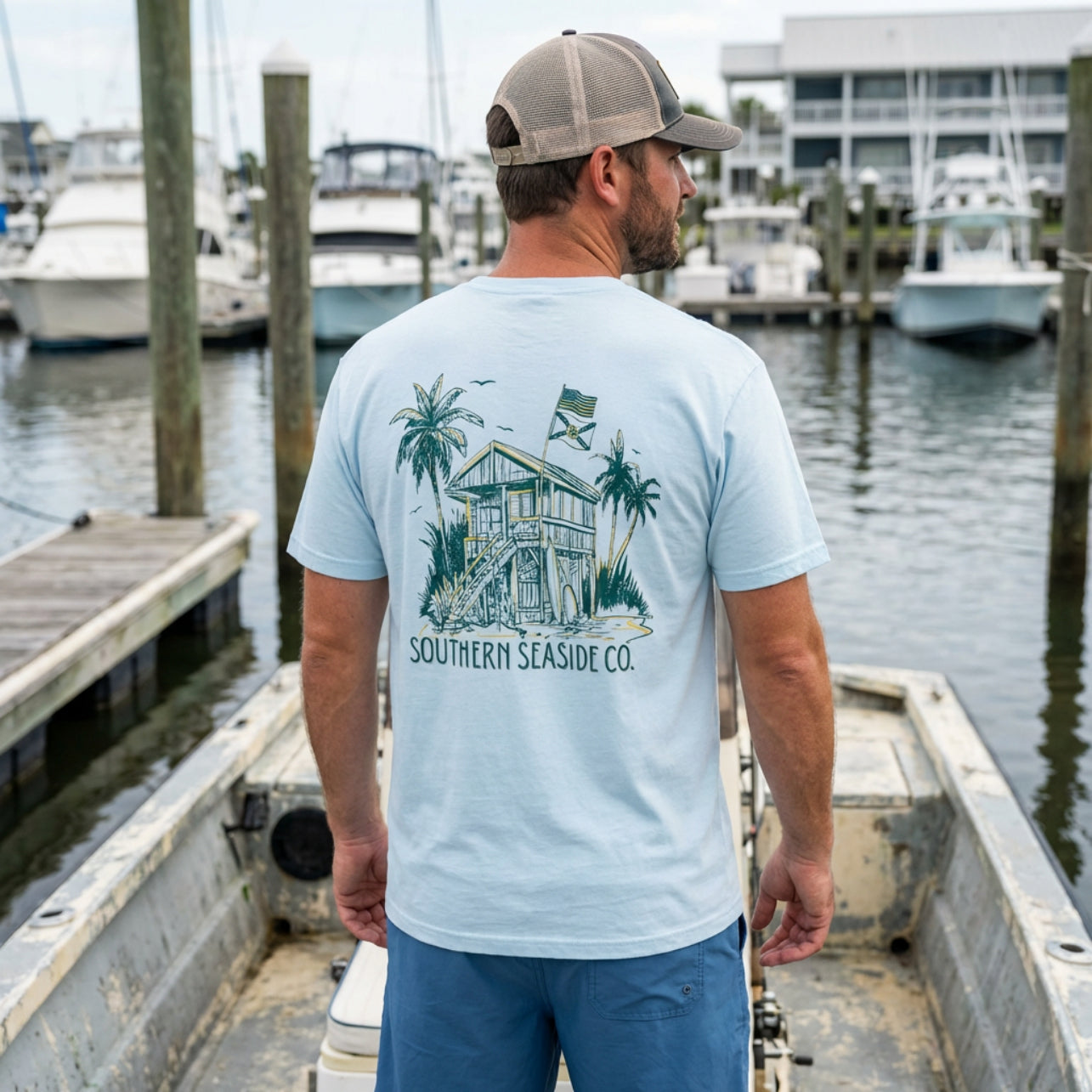 Man wearing a light blue t-shirt with a graphic design standing on a dock with boats in the background.