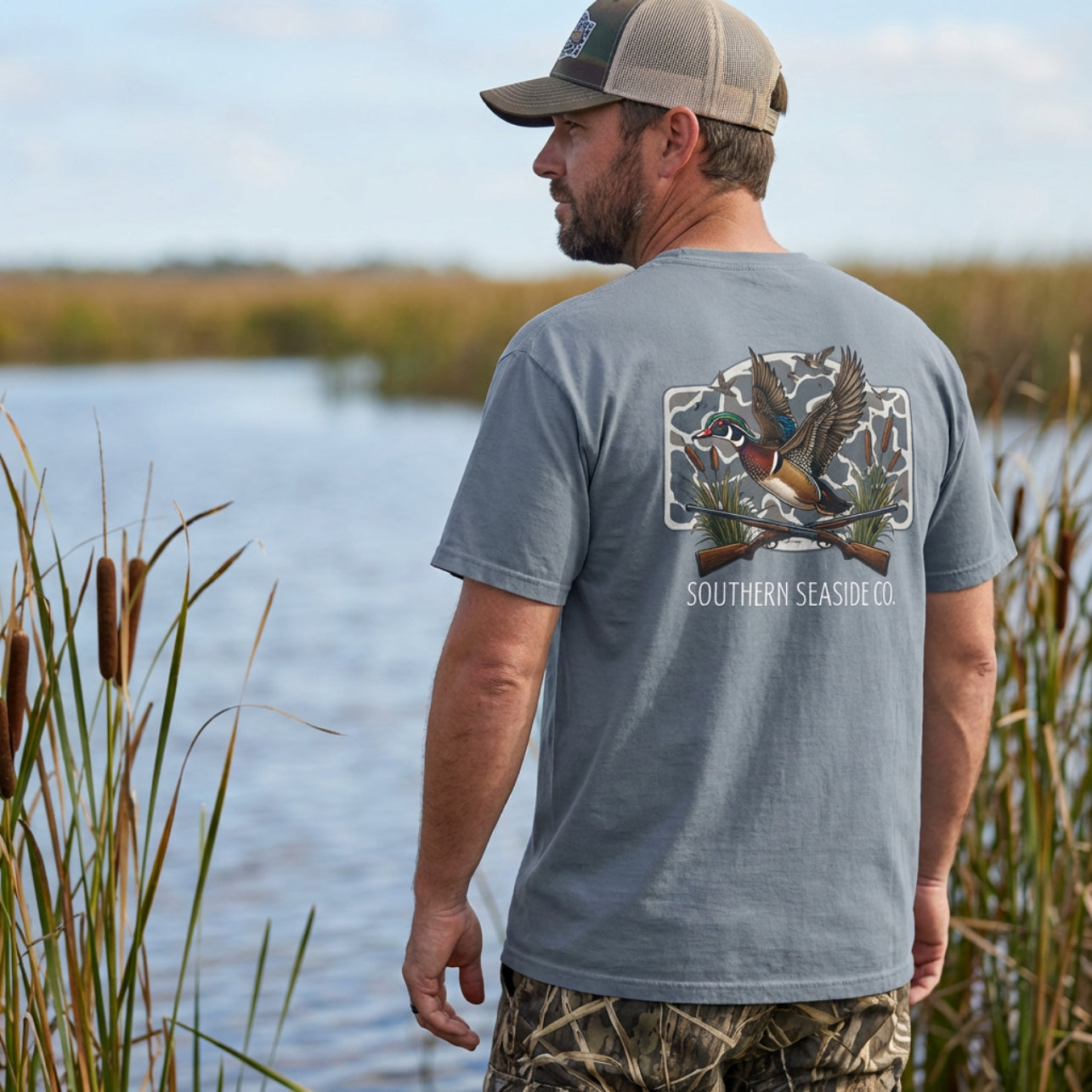 Man wearing a duck hunting gray t-shirt with a duck hunting and camouflage design standing by a body of water