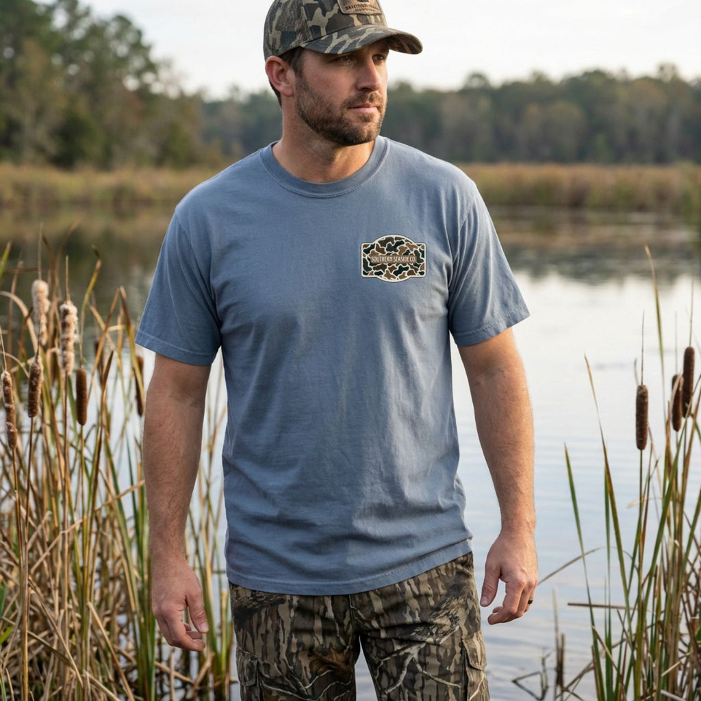 Man wearing a blue t-shirt with a logo and camouflage pants standing by a body of water with duck hunting tshirt and camo pants.