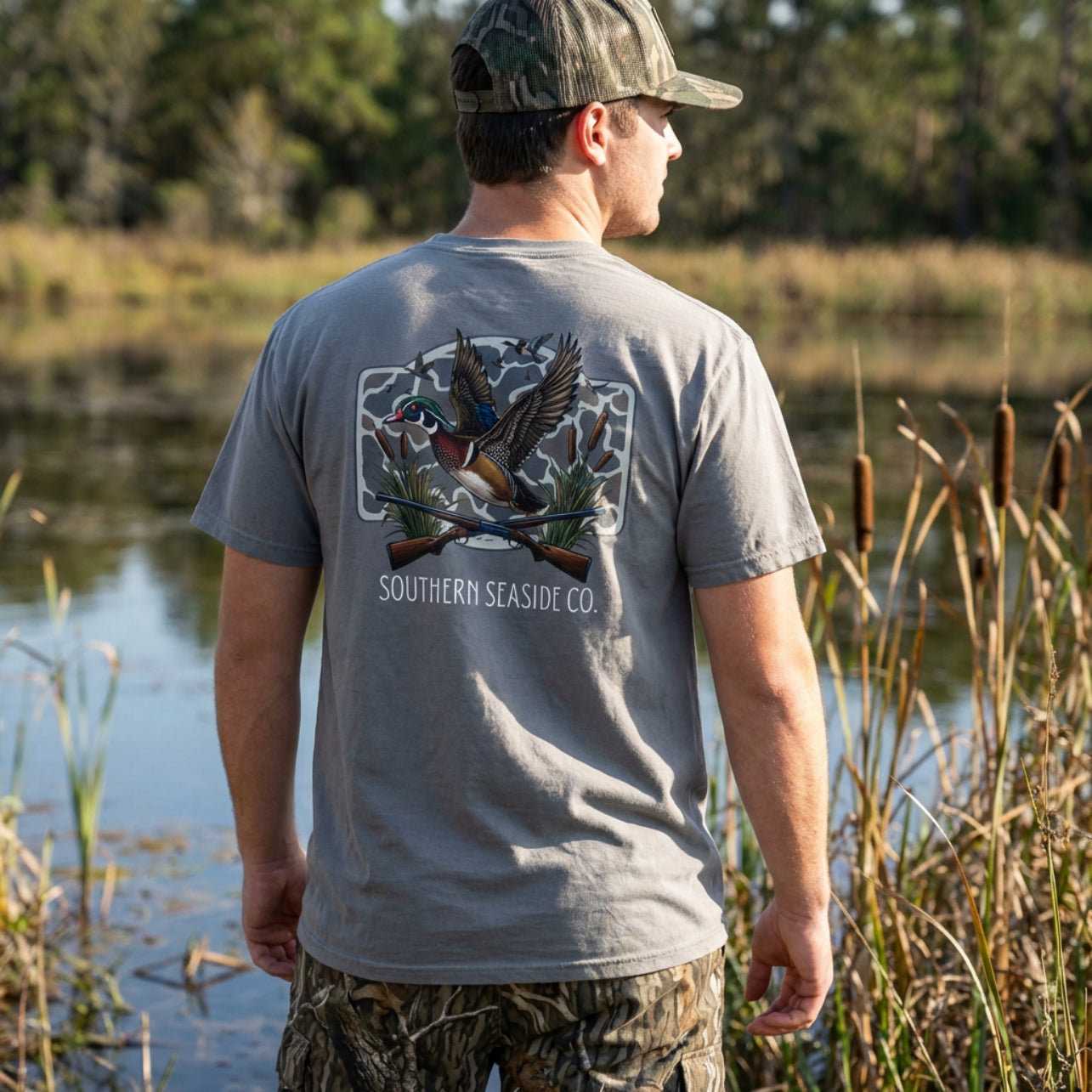 Man wearing a gray t-shirt with duck  hunting design and 'Southern Seaside Co' logo text, standing by a body of water duck hunting.