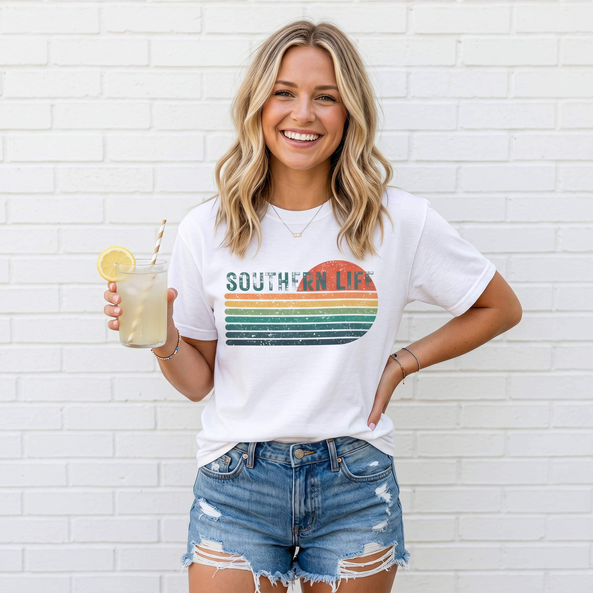 Woman wearing a 'Southern Life' t-shirt with a colorful design, holding a drink against a white brick wall.