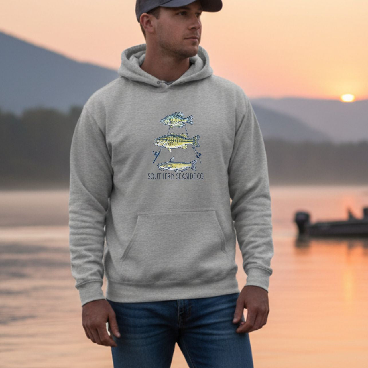 A man wearing a light gray Hanes Ultimate Cotton hoodie featuring a stacked triple‑fish graphic and Southern Seaside Co. logo, standing outdoors near the water at sunset with mountains and a boat in the background